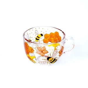 Op de afbeelding: Een heldere glazen theekop met een handvat, versierd met handgeschilderde bijen, honingraat en bloemen. De bijen zijn geel en zwart, de honingraat is oranje en de bloemen zijn oranje en bruin. De beker is voor thee of koffie.