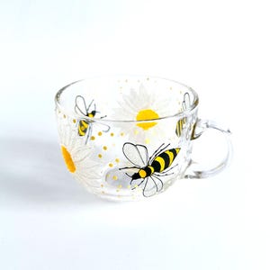 May include: A clear glass mug featuring a hand-painted design of bees and daisies. The bees are yellow and black, and the daisies are white with yellow centres.