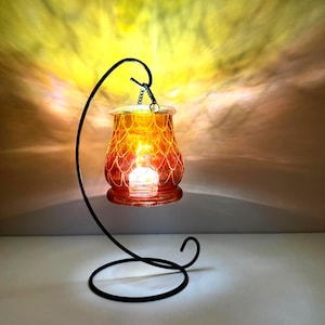 Könnte beinhalten: Ein Glaskerzenhalter mit orange-gelbem Farbverlauf und weißem Schuppenmuster, der an einem geschwungenen schwarzen Metallständer hängt. Der Halter ist von innen beleuchtet und wirft einen warmen Schein sowie Lichtmuster auf die Umgebung.