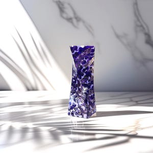 May include: A decorative vase with a distinctive, twisted shape. The vase showcases a rich pattern of deep purple and gold, offering a striking visual appeal. The glossy material of the vase reflects light, and it stands on a white surface, with shadows adding depth.