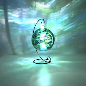 Op de afbeelding: Een decoratieve glazen bol met blauwe en groene wervelingen, van binnenuit verlicht. De bol wordt ondersteund door een zwarte metalen standaard met een gebogen ontwerp. De achtergrond toont een wazig, abstract patroon in blauw- en groentinten.