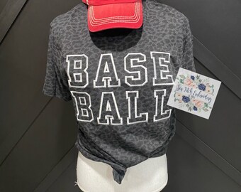May include: A black and gray leopard print t-shirt with the word "BASEBALL" in white letters. The shirt is on a white mannequin. A red baseball cap with the words "WORLD SERIES" and "BASEBALL" is on top of the mannequin's head.