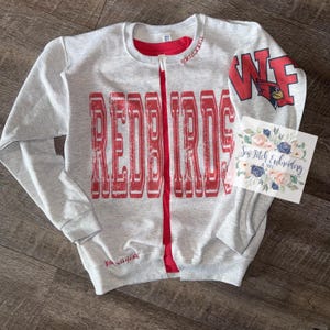 May include: A light gray zip-up sweatshirt with the word "REDBIRDS" in distressed red lettering across the front. The sweatshirt has a red zipper and a red lining. The right sleeve features "WF" and a cardinal logo. A small floral card is on the right.