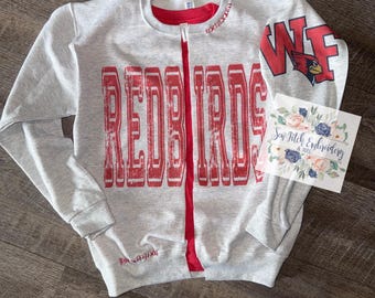 May include: A light gray zip-up sweatshirt with the word "REDBIRDS" in distressed red lettering across the front. The sweatshirt has a red zipper and a red lining. The right sleeve features "WF" and a cardinal logo. A small floral card is on the right.