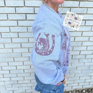 K&ouml;nnte beinhalten: Ein hellgraues Sweatshirt mit einem weinroten Hufeisen und einem Rodeo-Motiv auf dem &Auml;rmel. Das Sweatshirt hat auch eine gr&ouml;&szlig;ere Grafik auf der Vorderseite. Der Text "Sew Stitch Embroidery" ist auf einem kleinen Aufkleber zu sehen.