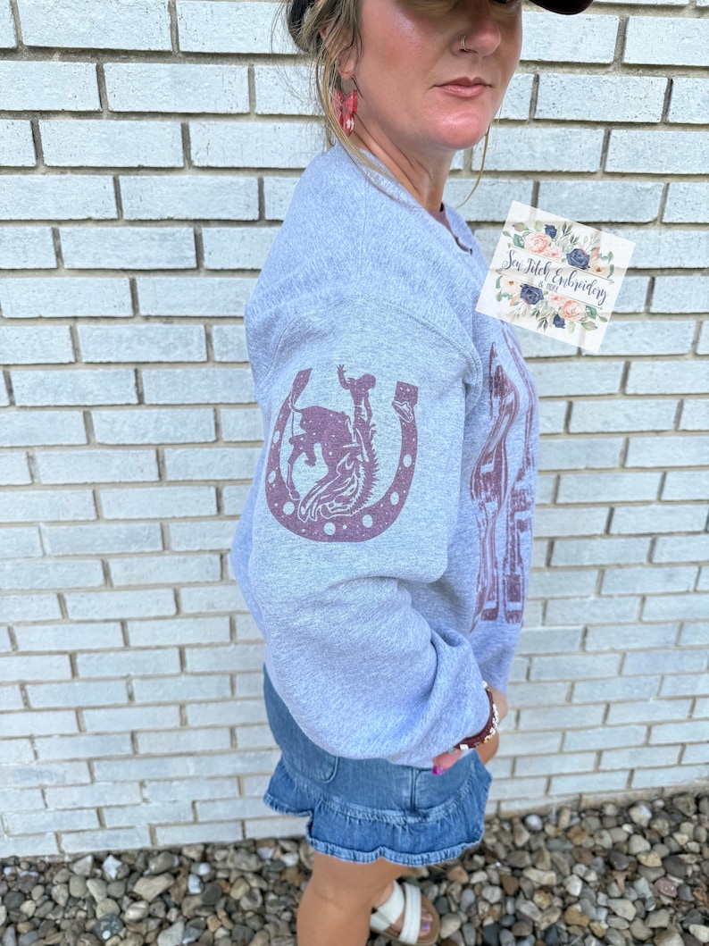 K&ouml;nnte beinhalten: Ein hellgraues Sweatshirt mit einem weinroten Hufeisen und einem Rodeo-Motiv auf dem &Auml;rmel. Das Sweatshirt hat auch eine gr&ouml;&szlig;ere Grafik auf der Vorderseite. Der Text "Sew Stitch Embroidery" ist auf einem kleinen Aufkleber zu sehen.