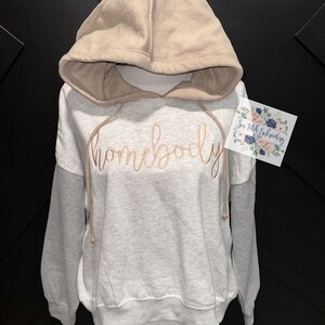 May include: A gray and beige hoodie with a tan hood and the word "homebody" embroidered in gold on the front. The hoodie is on a white mannequin.