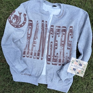 K&ouml;nnte beinhalten: Ein graues Sweatshirt mit Rei&szlig;verschluss und dem Wort "RANGERS" in verblichener brauner Schrift auf der Vorderseite. Auf dem linken &Auml;rmel befindet sich eine Hufeisen- und Cowboy-Grafik. Das Sweatshirt hat ein wei&szlig;es Innenfutter. Ein kleines Etikett sagt "Sew Stitch Embroidery".