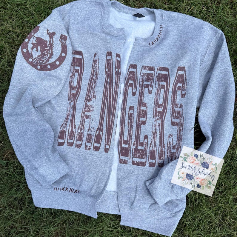 K&ouml;nnte beinhalten: Ein graues Sweatshirt mit Rei&szlig;verschluss und dem Wort "RANGERS" in verblichener brauner Schrift auf der Vorderseite. Auf dem linken &Auml;rmel befindet sich eine Hufeisen- und Cowboy-Grafik. Das Sweatshirt hat ein wei&szlig;es Innenfutter. Ein kleines Etikett sagt "Sew Stitch Embroidery".