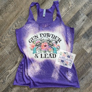 May include: Purple racerback tank top with a bleached design. The front features the text "GUN POWDER & LEAD" with crossed revolvers and floral accents. A small card with floral design is on the right.