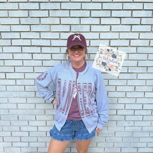 May include: A person wearing a gray zip-up jacket with the word "DANCER" printed in maroon. They are also wearing a maroon baseball cap with a white bow design, a maroon t-shirt, and denim shorts. A sign with floral art is held.