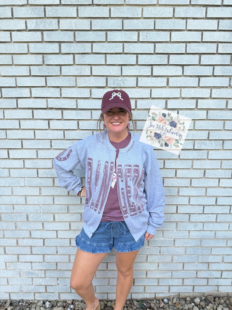 K&ouml;nnte beinhalten: Eine Person tr&auml;gt eine graue Rei&szlig;verschlussjacke mit dem Wort "DANCER" in Burgunderrot. Sie tr&auml;gt auch eine bordeauxfarbene Baseballkappe mit einem wei&szlig;en Schleifendesign, ein bordeauxfarbenes T-Shirt und Jeansshorts. Ein Schild mit floraler Kunst wird gehalten.