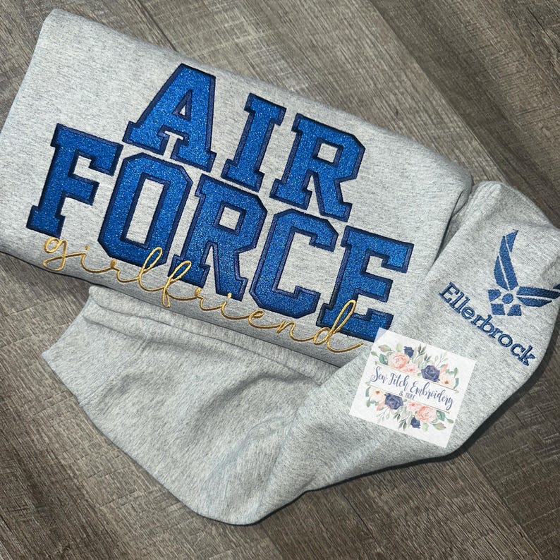 May include: Gray sweatshirt with blue glitter "AIR FORCE girlfriend" text and a blue Air Force logo on the sleeve. The sweatshirt is folded and a small white tag with the text "Sew Stitch Embroidery" is visible.