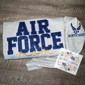 May include: Gray sweatshirt with blue glitter "AIR FORCE mom" text and a blue Air Force logo on the sleeve. The text "MONTGOMERY" is below the logo.