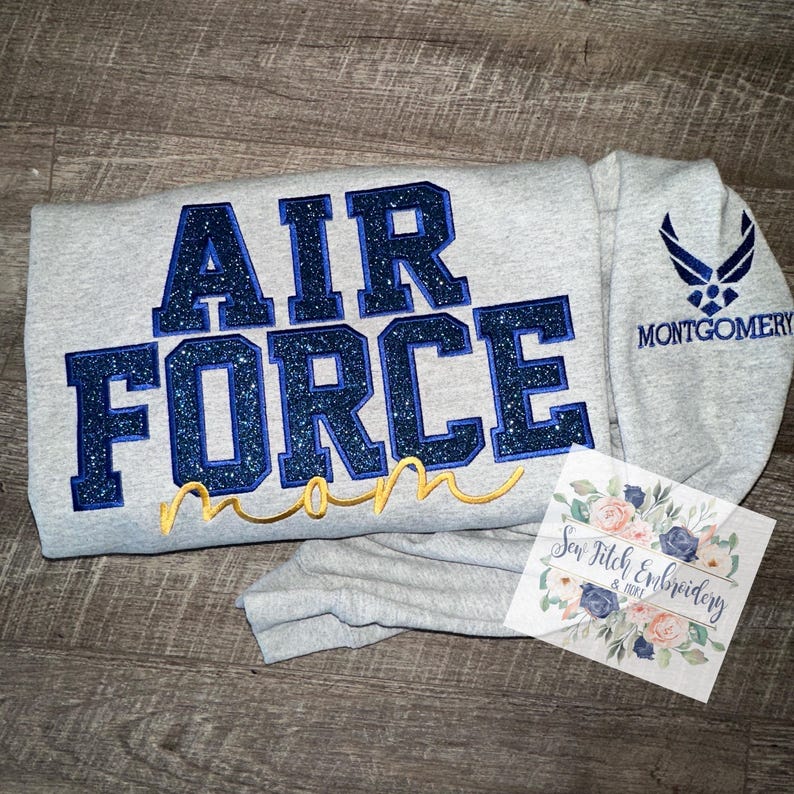 May include: Gray sweatshirt with blue glitter "AIR FORCE mom" text and a blue Air Force logo on the sleeve. The text "MONTGOMERY" is below the logo.