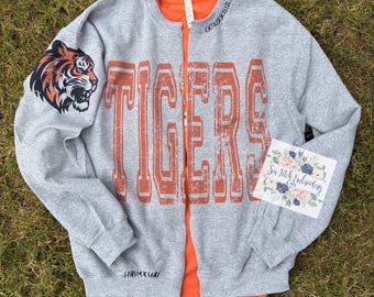 May include: A gray sweatshirt with the word "TIGERS" in orange block letters. A tiger head graphic is on the left sleeve. The sweatshirt is open, revealing an orange shirt underneath. A small floral patch is on the right side.