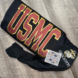 May include: Black long-sleeve shirt with a red and gold glitter "USMC" design on the front and a white embroidered USMC logo on the sleeve. The shirt also has the text "GIGLIOTTI" embroidered on the sleeve.