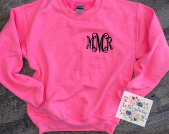 May include: A bright pink sweatshirt with a black embroidered monogram "MMCR" on the front. The sweatshirt is made of a soft, comfortable material and has a crew neck.