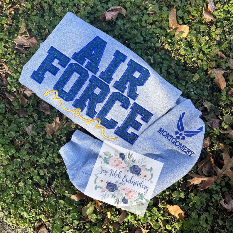 May include: Gray sweatshirt with blue glitter embroidery that reads "Air Force Mom" and a blue Air Force logo on the sleeve. The sweatshirt is laying on a bed of green grass.