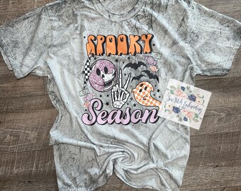 May include: A white t-shirt with a distressed, tie-dye effect. The shirt features a graphic design with the text "Spooky Season" in pink and orange letters. The graphic also includes a skull, a ghost, and bats.