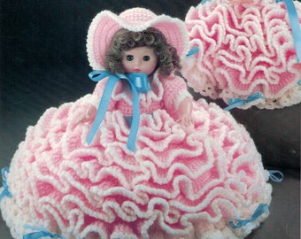 May include: Two crocheted pillow dolls with ruffled pink and white skirts. The dolls are wearing blue ribbons and hats. The text on the image reads "10 1/2" Pillow Doll..... Half doll body attaches to a crocheted pillow base to create a firmly stuffed skirt. Also includes directions for 13" Bed Doll dress."