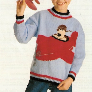 Könnte beinhalten: Hellblauer Strickpullover mit rotem Flugzeugdesign auf der Vorderseite. Der Pullover hat schwarze und rote Verzierungen an Kragen, Manschetten und Saum. Ein Kind hält einen roten Flugzeug-Ausschnitt.