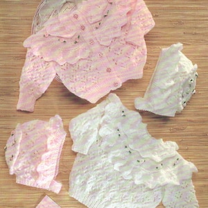 May include: Two knitted baby sweaters with ruffled edges and floral embellishments, one pink and one white, and two matching knitted hats with ruffled edges.
