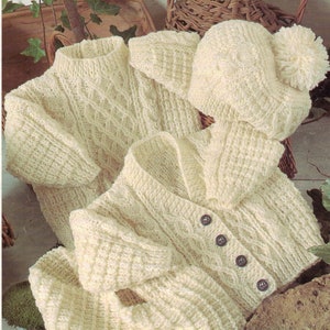 Puede incluir: Un conjunto de punto blanco para bebé que incluye un cárdigan, un gorro, un jersey y una bufanda. El conjunto presenta un patrón de punto de cable.