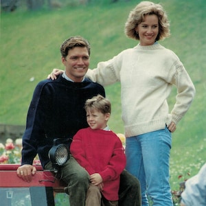 Könnte beinhalten: Eine Familie posiert im Freien. Der Mann trägt einen marineblauen Pullover und eine grüne Hose. Die Frau trägt einen cremefarbenen Pullover und blaue Jeans. Ein Kind in einem roten Pullover sitzt zwischen ihnen. Der Hintergrund ist grün.