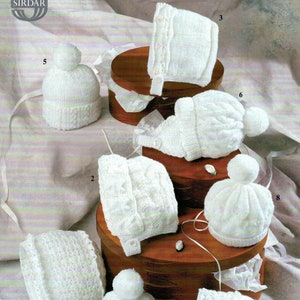 Op de afbeelding: Tien witte gebreide baby mutsen met verschillende ontwerpen en stijlen. Sommige mutsen hebben pompoenen, sommige hebben bandjes en sommige hebben kantversiering. De mutsen worden gepresenteerd op een houten oppervlak.