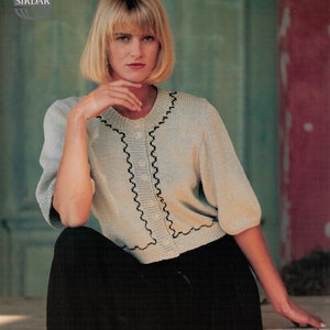 Puede incluir: Una mujer lleva un cárdigan de punto beige claro con ribetes negros y una falda negra. El cárdigan tiene cierre de botón y un ribete festoneado.