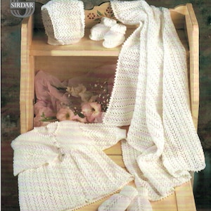 Può includere: Un set da neonato in maglia bianca con un motivo a pizzo. Il set comprende un cappello, scarpine, un cardigan e uno scialle. Gli articoli sono esposti su uno scaffale in legno con un cassetto.