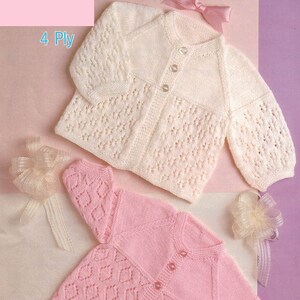 May include: Two knitted baby cardigans, one white and one pink, with a button closure and a delicate lace pattern. The cardigans are laid out on a purple background. 4 Ply.