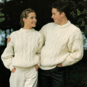 May include: Two people wearing white cable knit sweaters. The woman is wearing a brown headband and white pants. The man is wearing blue jeans.