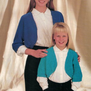 Op de afbeelding: Twee vrouwen dragen witte blouses en blauwe en turquoise gebreide bolero's. De vrouw vooraan draagt een zwarte rok en de vrouw achteraan draagt een zwarte rok en een zwart hoofdband.