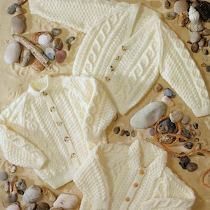 Op de afbeelding: Crèmekleurige gebreide vesten met kabelpatroon en knoopsluitingen. De truien worden getoond op een zanderige ondergrond met schelpen en kiezelstenen, waarbij de textuur en het ontwerp van de kledingstukken worden benadrukt.