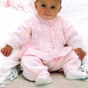 May include: A pink knitted baby cardigan and trousers set with a button fastening. The cardigan features a delicate lace pattern and the trousers have a frilled hem.