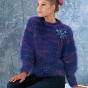 May include: A fuzzy, long-sleeved sweater in shades of blue and purple, with a floral embellishment on the chest. The sweater has a round neckline and a soft, textured appearance. The model is wearing dark blue jeans and a pink bow.