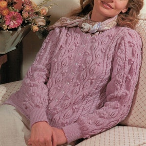 May include: A pink knitted cardigan with a cable knit pattern and pearl-like buttons. The cardigan is worn by a woman sitting on a couch.