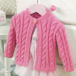 May include: A pink cable-knit cardigan with a single button closure, hanging on a pink hanger. The sweater has a round neckline and long sleeves. A white dress with floral embroidery is visible beneath the cardigan.
