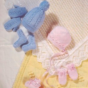 Puede incluir: Un conjunto de ropa de bebé de punto en azul y rosa. El conjunto incluye un gorro, botines y manoplas. Los artículos están dispuestos sobre una manta de ganchillo amarilla y blanca.