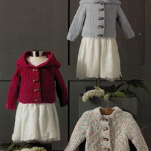 Pode incluir: Três cardigans infantis em exposição. Um é um cardigan com capuz vermelho escuro com botões de madeira, outro é um cardigan com capuz azul claro e o terceiro é um cardigan multicolorido com gola. Todos são combinados com saias brancas.