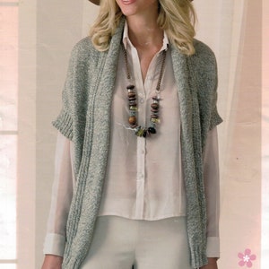 May include: A gray knitted cardigan with a wide collar and short sleeves. The cardigan is worn over a white button-down shirt and beige pants. The model is wearing a brown hat and a beaded necklace.
