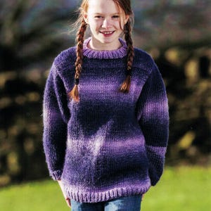 May include: A knitted purple sweater with a round neckline and long sleeves. The sweater features a striped pattern in shades of purple and blue. The person is wearing blue jeans.