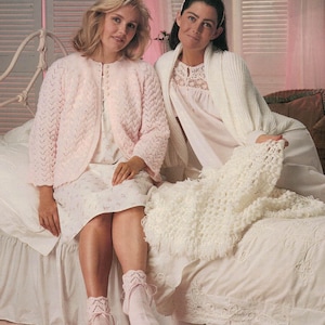 Könnte beinhalten: Zwei Frauen in passender pastellrosa Nachtwäsche sitzen auf einem Bett. Eine trägt eine Strickjacke, die andere einen weißen Bademantel. Eine weiße Häkeldecke liegt auf dem Bett. Der Raum hat eine weiche, feminine Ästhetik.