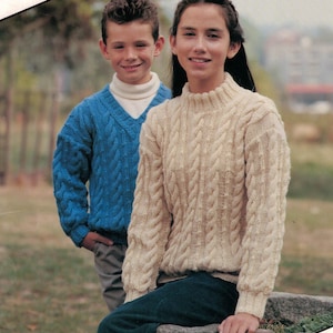 Puede incluir: Dos personas con suéteres de punto trenzado. Uno es azul claro con cuello en V, el otro crema con cuello redondo. Los suéteres tienen un diseño trenzado texturizado. La persona con el suéter crema está sentada en una piedra.