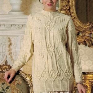 May include: A cream-coloured, long-sleeved sweater with a high neckline. The sweater has a detailed cable-knit design with diamond and leaf patterns. The model is wearing a floral skirt and standing in a room with ornate decor.