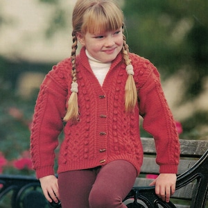 Puede incluir: Una persona joven con una rebeca roja de punto con botones de madera, un cuello alto blanco y leggings burdeos. La rebeca presenta un patrón texturizado. La persona tiene trenzas rubias y está sonriendo.