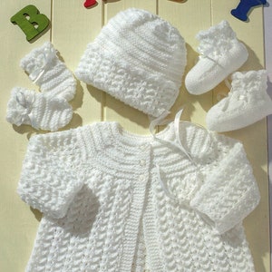 May include: A white knitted baby outfit set, including a cardigan, hat, and booties. The set is laid out on a light wood background with the word "BABY" spelled out in colorful letters.
