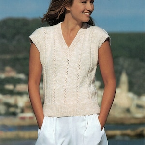 以下が含まれることがあります： Vネックと繊細な透かし模様が施された白いノースリーブニットベスト。長いブロンドの髪をした人が、海岸の町のぼやけた背景に立ってベストを着ています。